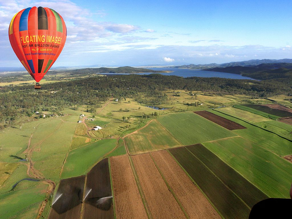 Floating Images Hot Air Balloon Flights - Red hot air balloon with coloured stripes at the top, floating over the green countryside of Somerset with views of Lake Wivenhoe in the distance.