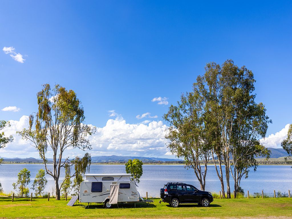 Caravan set up behind a car at the NRMA Lake Somerset Holiday Park beside the lake. It is a sunny day with blue sky, green grass and a couple of gum trees either side of the caravan.
