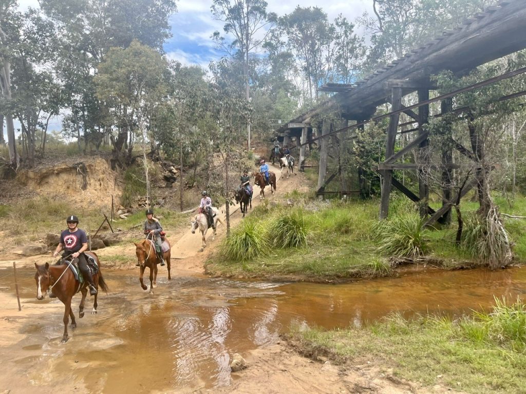 A line of horse riders crossing a shallow creek in single file along the Brisbane Valley Rail Trail, surrounded by Australian bush. An old wooden railway bridge is in the background on the right side.