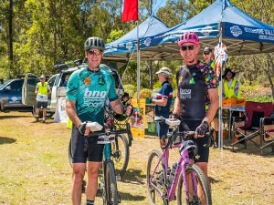 Two male cyclists posing for a photo with their bikes infront of the blue Brisbane Valley Rail Trail Users Association Tent after a riding event