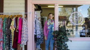 Little Country Lane shopfront. Female owner standing in the pink doorway of the shop with a rack of clothes out the front, and plants outside the doorway.