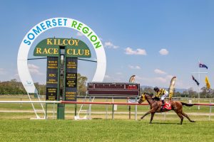 Kilcoy Races - horse and jockey crossing the finish line at the Kilcoy Showground. Green grass on the track and blue sky.