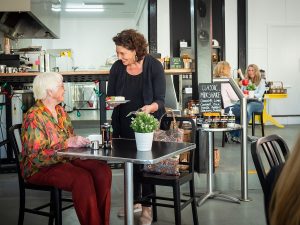 An older lady enjoying a conversation at the Picnic Society when being served her order. People seated in the background of the cafe.