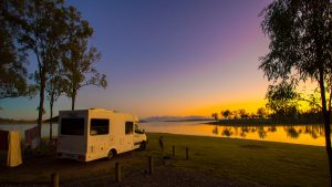 Campervan parked beside Lake Wivenhoe. The photo was taken at sunset with the sky a mix of orange, and purple above the lake. Gum trees can be seen beside the campervan.