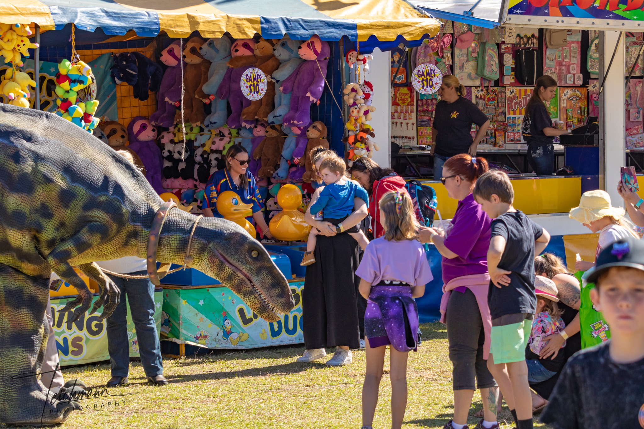 Sideshow Alley at the Esk Show. Kids and parents looking at and playing sideshow games. A group of kids are looking at a robotic dinosaur