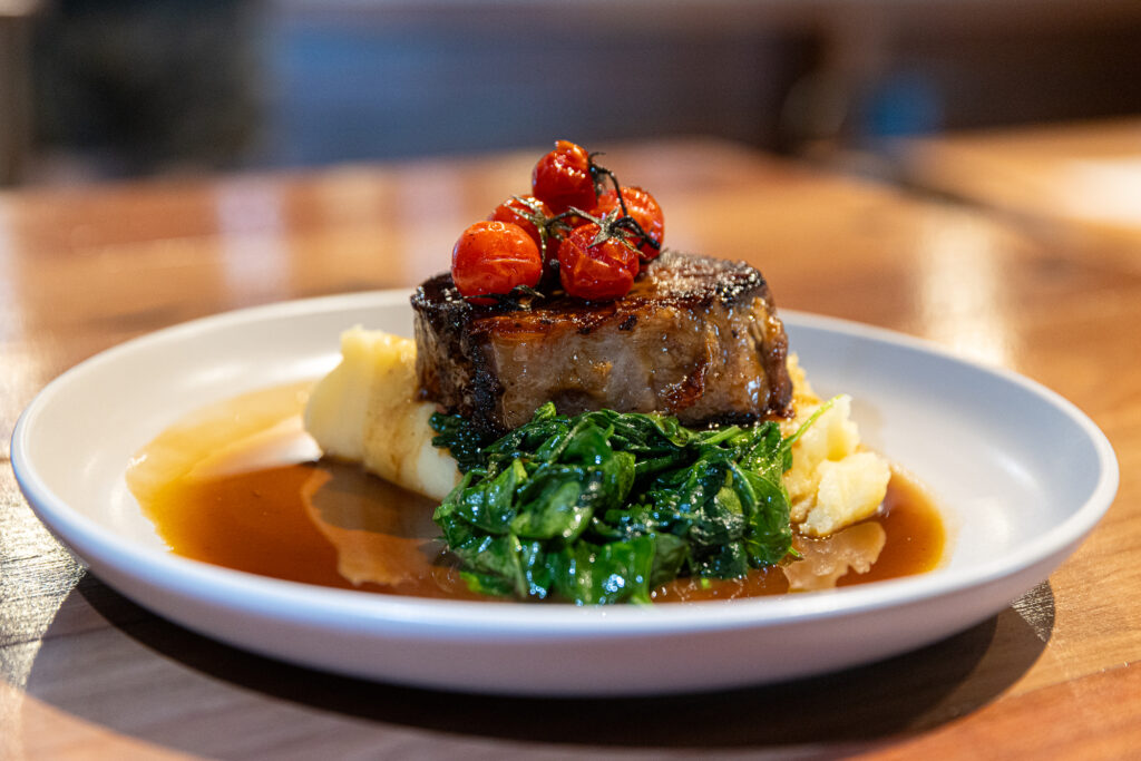 Splityard Social signature Beef Dish. White plate on a wooden table featuring Beef dish with gravy, mashed potato, greens, beef and tomatoes.