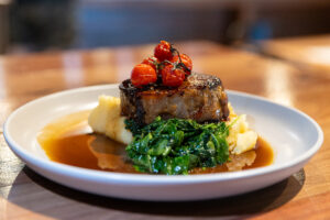 Splityard Social signature Beef Dish. White plate on a wooden table featuring Beef dish with gravy, mashed potato, greens, beef and tomatoes.