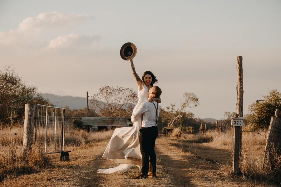 Groom holding up bride, waving a cowboy hat for a wedding photo pose.