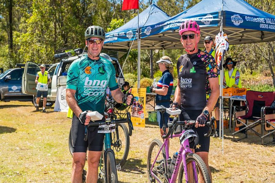 Two male cyclists posing for a photo with their bikes infront of the blue Brisbane Valley Rail Trail Users Association Tent after a riding event