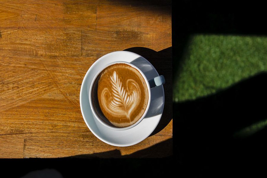 Overhead photo of a coffee with coffee art in the froth. The Coffee cup is sitting on top of a wooden table over a grassed area.