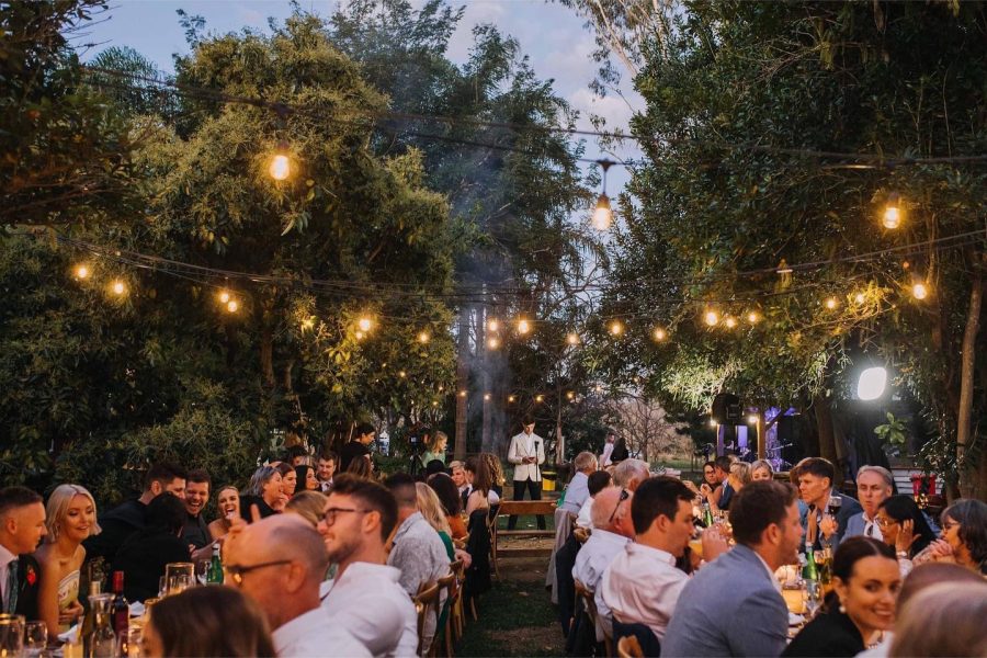 Wedding guests seated at outdoor tables. Fairy lights glowing above two rows of tables full of guests.