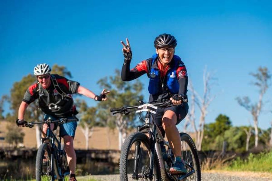 Two Cyclists riding and waving along the Brisbane Valley Rail Trail. Blue sky in the background along with the gravel trail and trees in the distance.