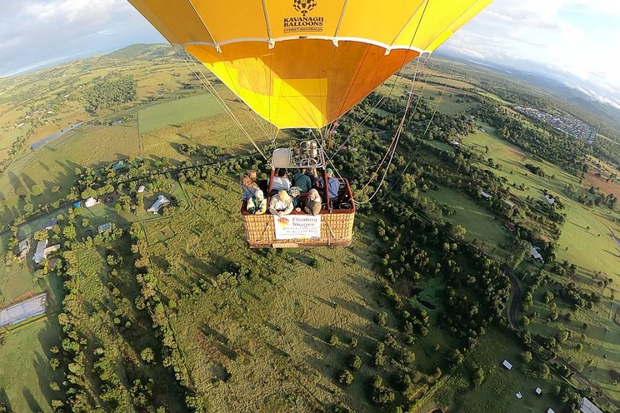 Close up of people in the basket of a yellow hot air balloon floating over the green countryside (taken via neighbouring balloon or drone).