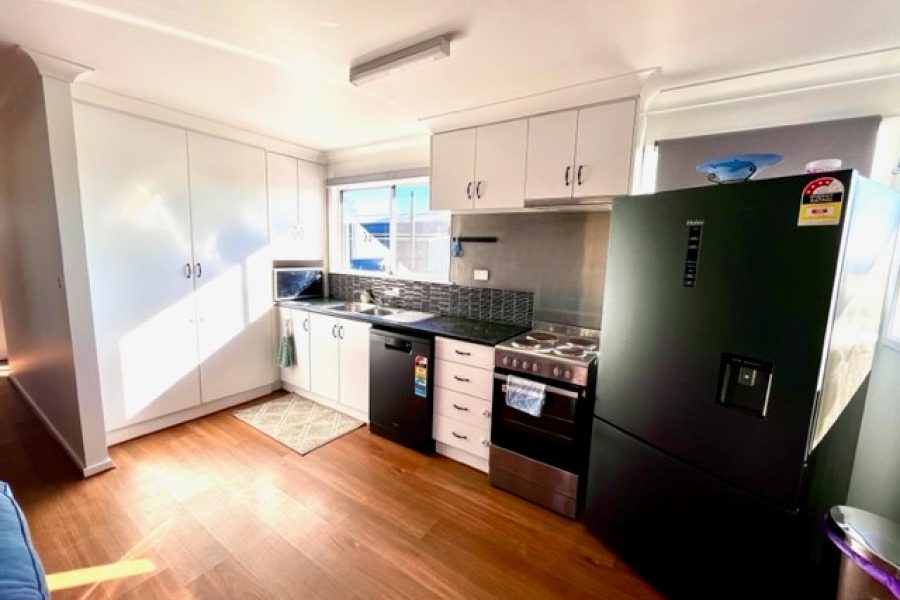 Photo of the kitchen inside Clarendon Cottage. Timber floors, white cabinets and counters, oven, and double door black fridge.