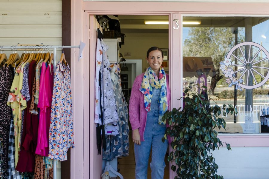 Little Country Lane shopfront. Female owner standing in the pink doorway of the shop with a rack of clothes out the front, and plants outside the doorway.