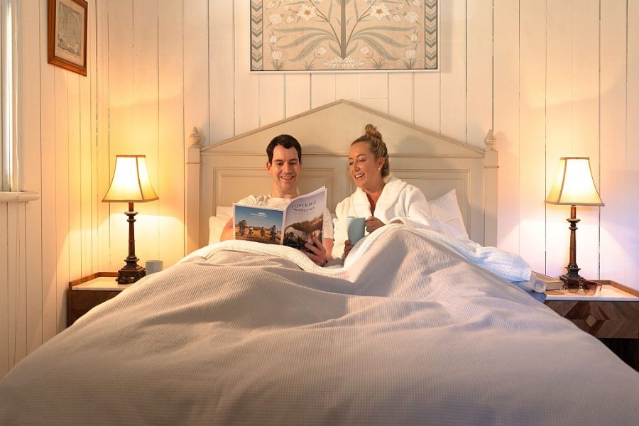 A couple are in bed at Montrose on Moore reading the Experience Somerset Visitor Guide. The room is soft pale colours, white timber walls, pale bedspread and bed side lamps are providing light.