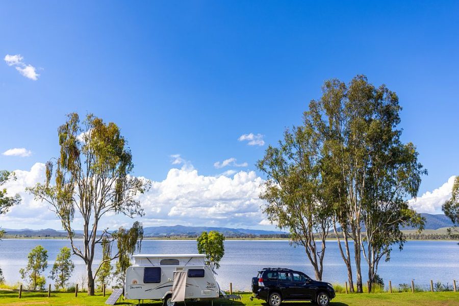 Caravan set up behind a car at the NRMA Lake Somerset Holiday Park beside the lake. It is a sunny day with blue sky, green grass and a couple of gum trees either side of the caravan.