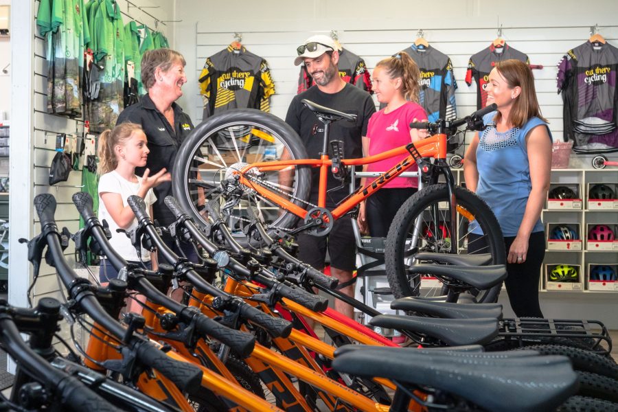 Family of four getting geared up inside the Brisbane Valley Rail Trail Cycles shop. The female shop owner is talking to the family, The shop has a range of mountain bikes on display with bike jerseys hanging on the wall behind the family.