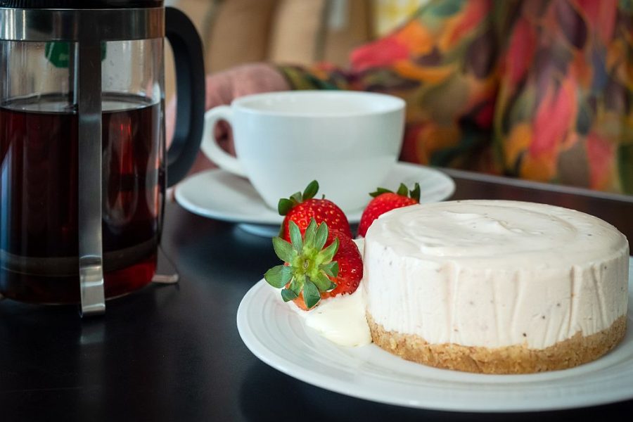 Close up photo of a Picnic Society Cheescake with a strawberry on the side. A white tea cup and black and glass teapot is in the background.