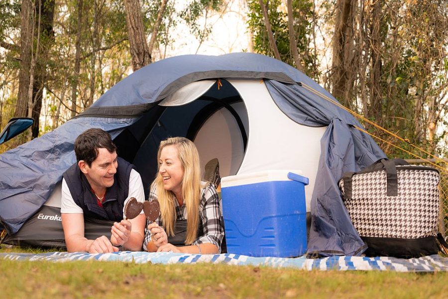 Couple leaning out of the pop up tent eating ice creams at Jimna Base Camp. Bushland setting behind them. Esky and picnic blanket in front of the blue and white tent.