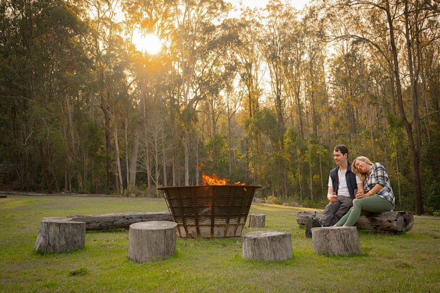 Couple sitting beside a campfire in a cast iron brazier on logs, at sunset. Tall gum trees surround them with a golden sunset glow.