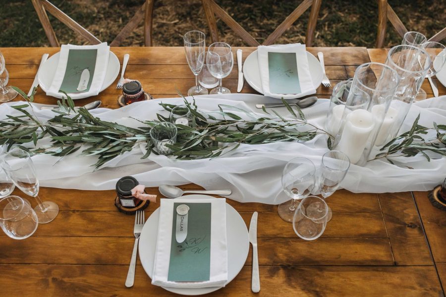 Wedding table setting. Timber table, with white flowing fabric as a table runner, white plates, glasses, cutlery and green branches as centre pieces.