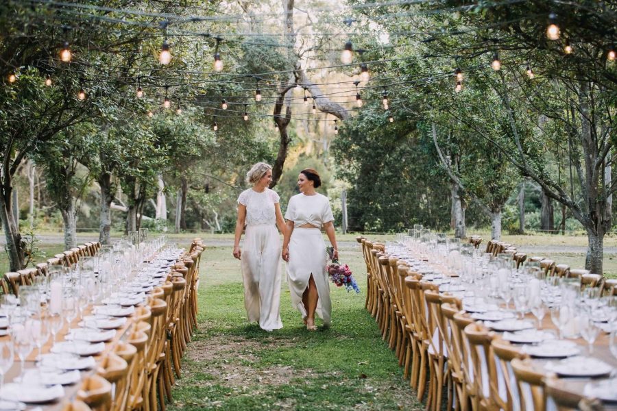 Two Brides dressed in white, walking down the aisle between the tables with fairy lights glowing above them.
