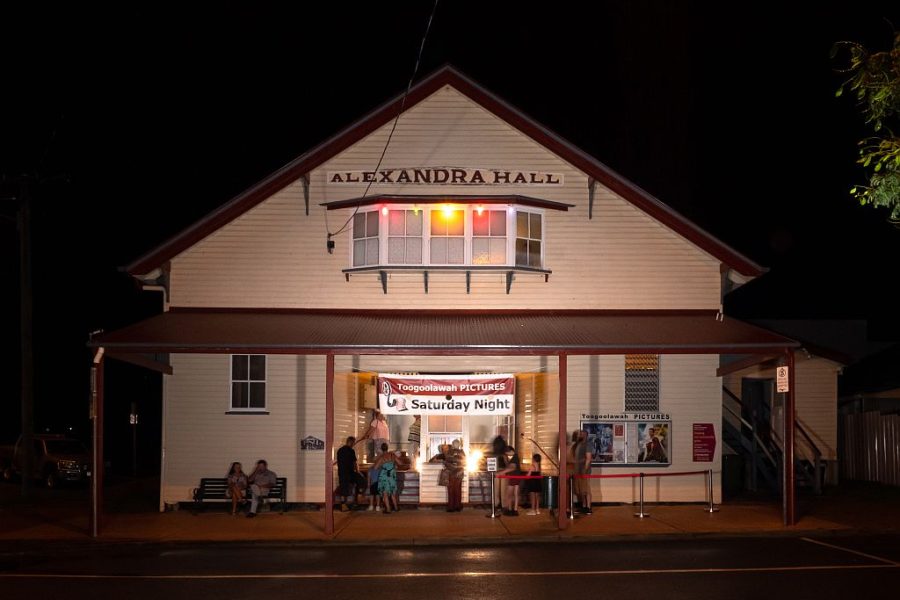 Outside the Toogoolawah Pictures at Alexandra Hall, An old style Hall with a box office out the front. Photo is taken at night with people waiting to buy tickets